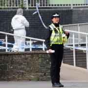 A police officer stands guard at Ysgol Dyffryn Aman in Ammanford where two adults and a child were stabbed, with a forensic officer in the background.